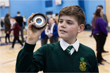 Young person in a school hall holding a piece of scientific equipment as other young people in the background work 