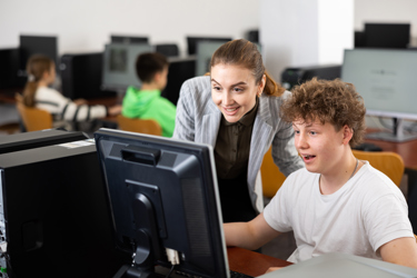 Young people in a school computer lab. A teacher stands with a student who's sitting at their desk looking at the computer screen.