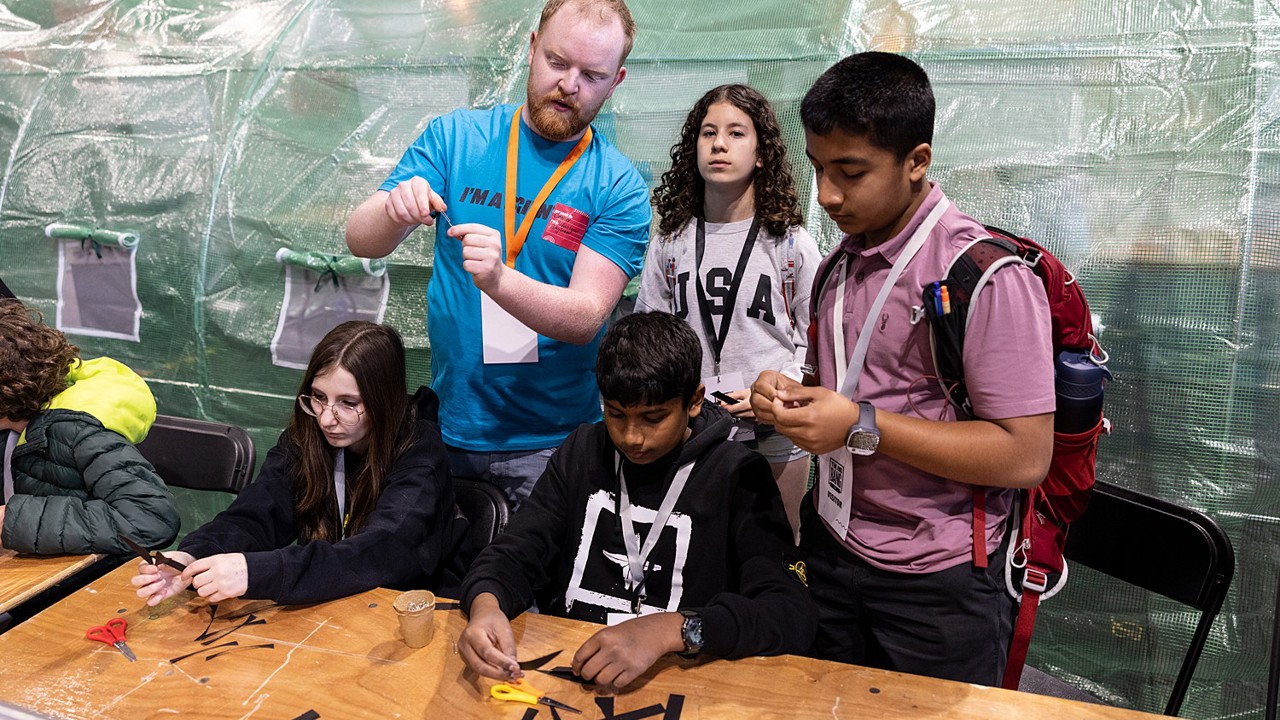 A volunteer at The Big Bang Fair demonstrates a task to four young people. Two of these people are sat at at table as they work on the task while the other two stand. All students are wearing lanyards.