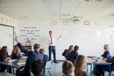 A teacher at the front of a class or secondary school students teaching. One student has their hand up