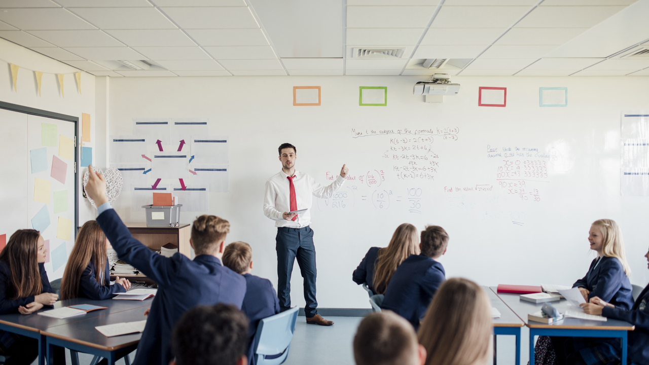 A teacher at the front of a class or secondary school students teaching. One student has their hand up