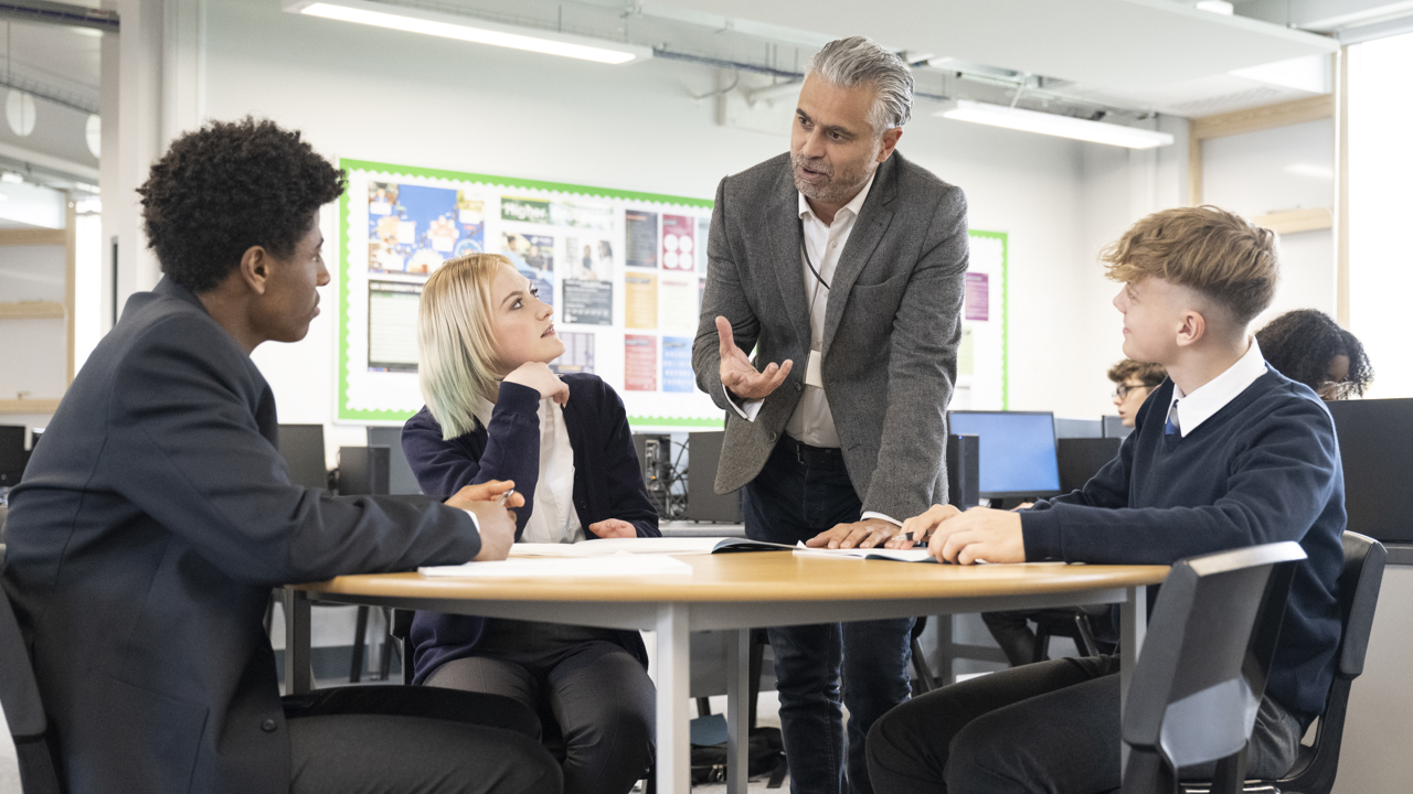 School classroom setting, three young people at a desk, a teacher bends to chat to them 