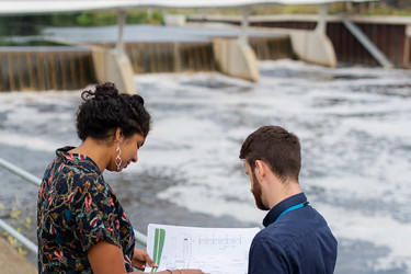 Two people stand by some water works looking at plans