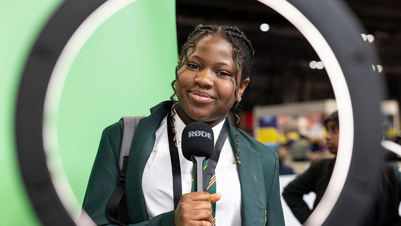 A young person at The Big Bang Fair holds a microphone ad looks through a ring lamp directly into camera