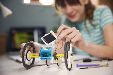 A young person has a small model of a solar panel on their desk along with other equipment such as pens and their notebook. They are holding the model. 