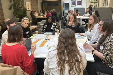 A working session in a meeting room, groups of people sit around tables discussing a topic