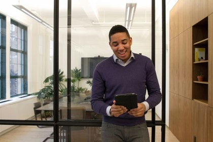 A young person stands in an office in front of an empty boardroom. They smile and look at the screen in their hands.