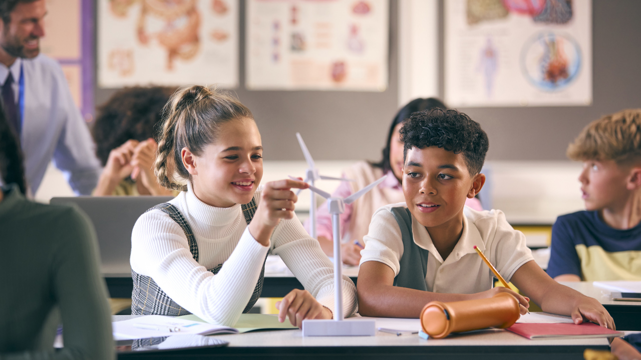 Primary school pupils in a classroom setting. The pupils have small model wind turbines on their desks along with text books and pencils. Two students are interacting with their model.
