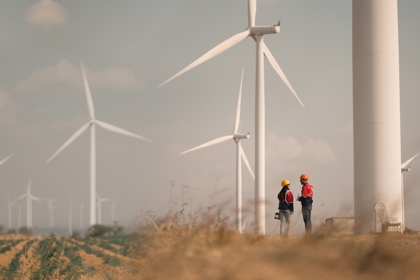 Two people in hard hats and protective clothing stand in a field of wind turbines. 