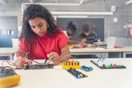 In the foreground a young person sits at a workbench looking at electronic circuit boars. Two people are doing the same in the background,